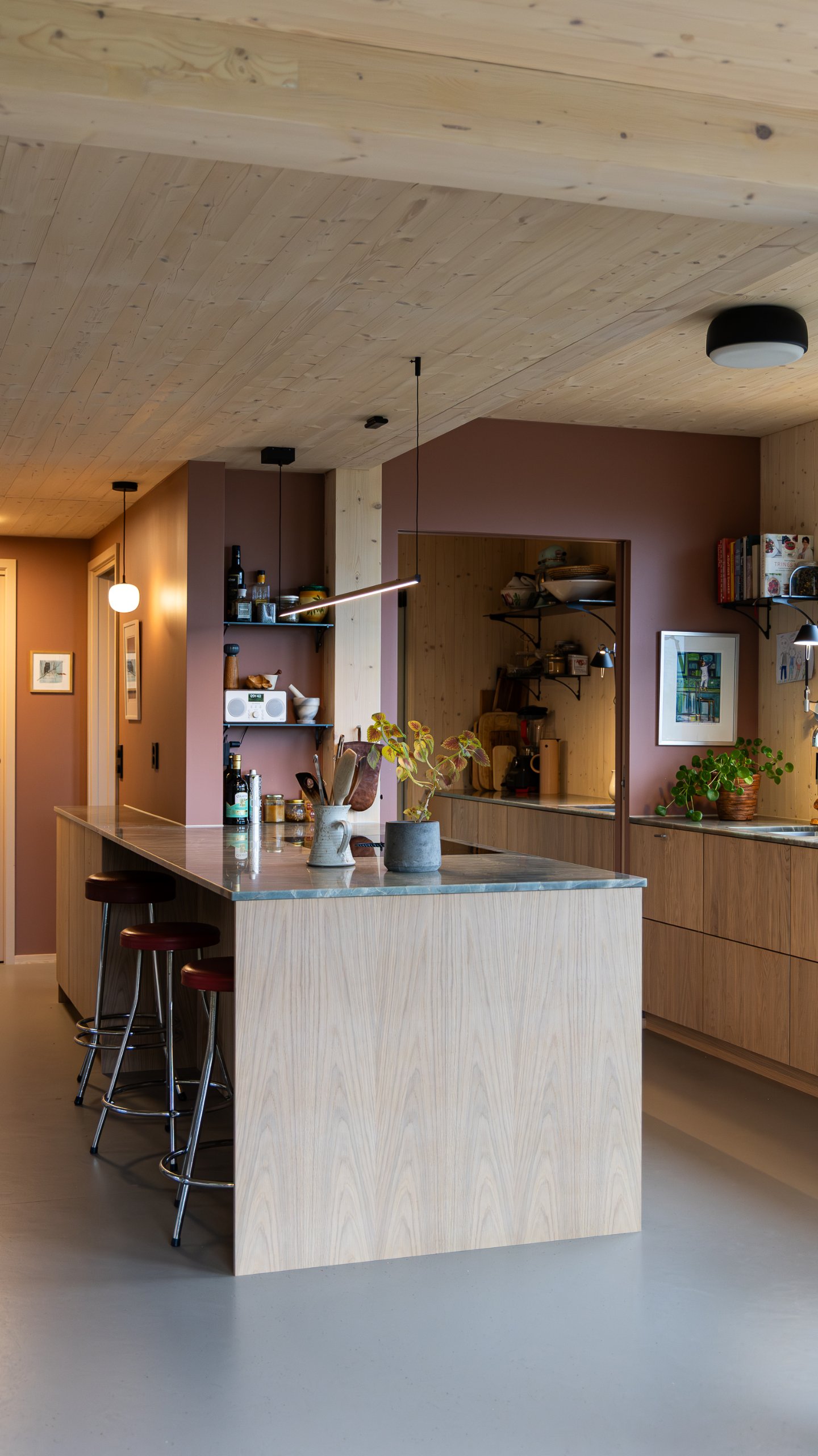 Kjøkkenøy med tre barkrakker under en smal pendellampe, omgitt av trepanel og åpne hyller. Kjøkkenøy med tre barkrakker under en smal pendellampe, omgitt av trepanel og åpne hyller.