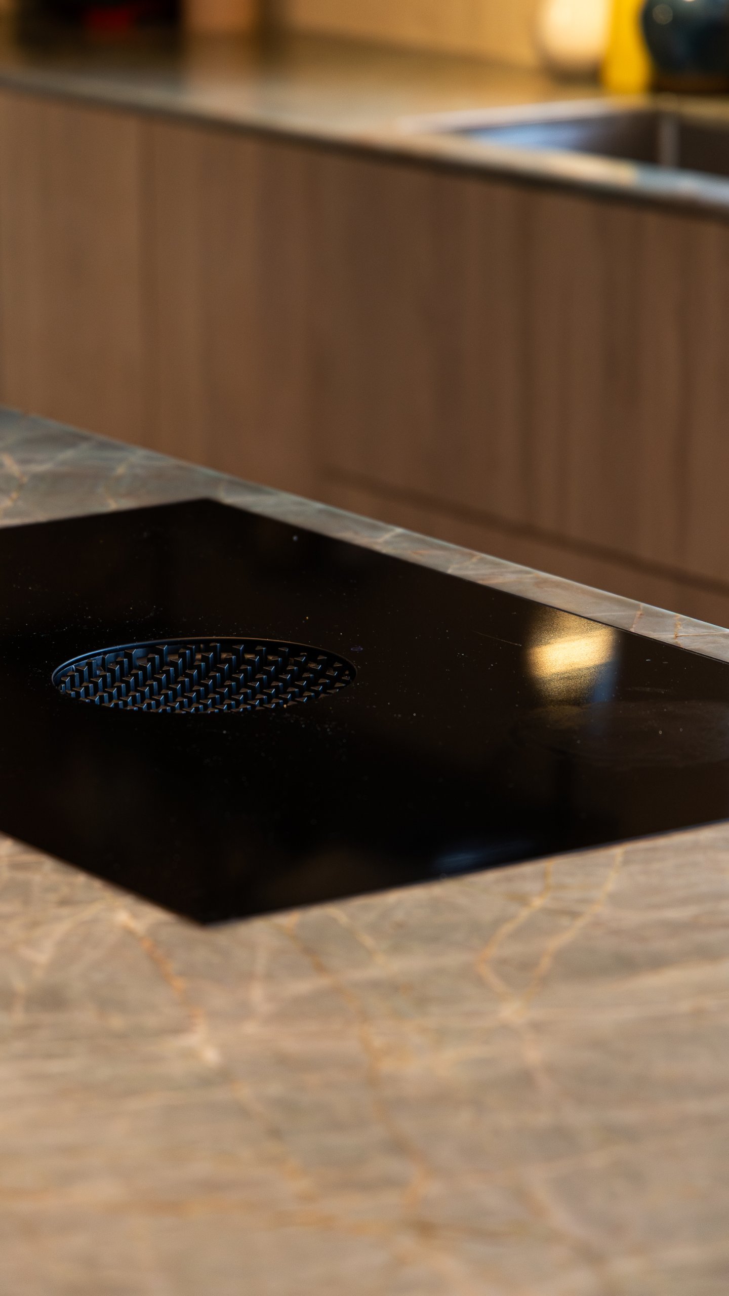 Nærbilde av svart platetopp med rundt ventilasjonsgitter, innfelt i kjøkkenøyas årede steinplate. Nærbilde av svart platetopp med rundt ventilasjonsgitter, innfelt i kjøkkenøyas årede steinplate.