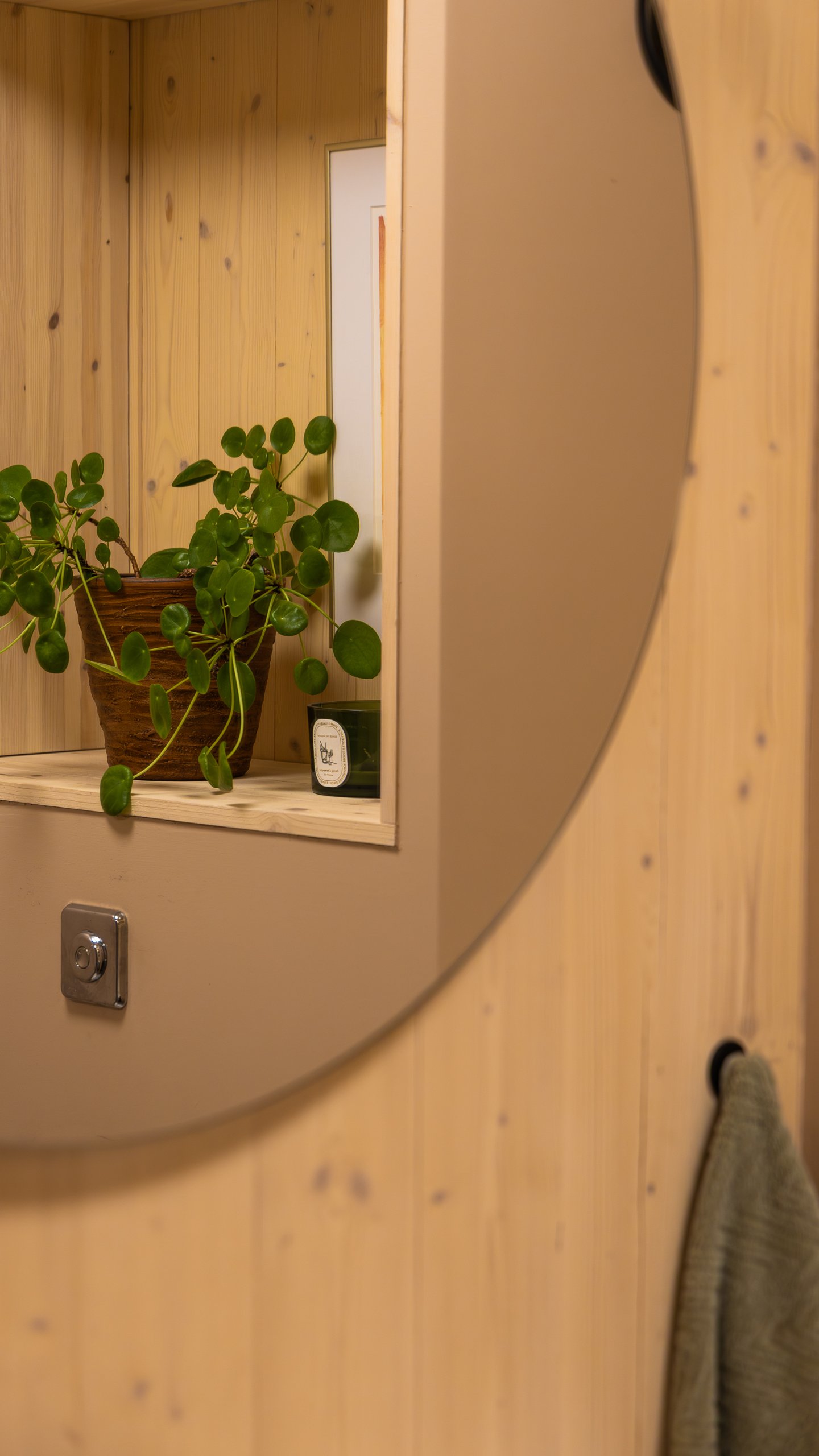 Rundt speil på trevegg med en nisje og potteplante i speilbildet samt et håndkle på krok ved siden av. Rundt speil på trevegg med en nisje og potteplante i speilbildet samt et håndkle på krok ved siden av.